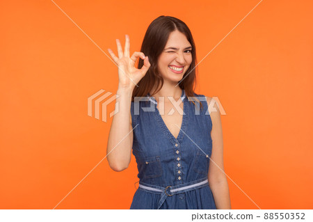 Okay, sure. Delighted happy brunette woman in denim dress smiling broadly, winking and showing ok gesture, approval sign, agreeing with suggestion. indoor studio shot isolated on orange background Okay, sure. Delighted happy brunette woman in denim dress smiling broadly, winking and showing ok gesture, approval sign, agreeing with suggestion. indoor studio shot isolated on orange background 88550352