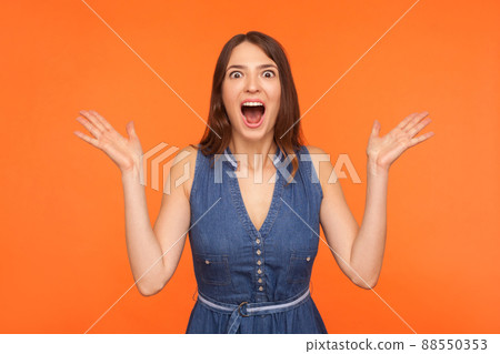 Shocked impressed brunette woman in denim outfit standing with wide open mouth and raising hands in surprise, looking startled amazed at camera. indoor studio shot isolated on orange background 88550353