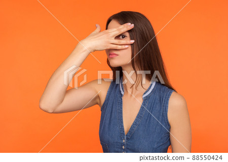 Curious nosy brunette woman in denim dress spying secrets, looking through fingers, peeking with interest at camera, feeling shy, shame to watch. indoor studio shot isolated on orange background 88550424