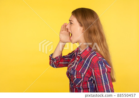Attention, advertising. Side view of girl in checkered shirt holding palm near mouth and shouting loudly, screaming, proclaiming message, crazy news. indoor studio shot isolated on yellow background 88550457