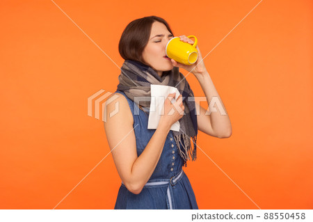 Flu treatment. Sick woman wrapped in scarf drinking tea and holding napkin for runny nose, treating influenza symptoms with hot beverage, feeling unwell. studio shot isolated on orange background 88550458