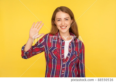 Hello. Portrait of kind friendly ginger girl in checkered shirt waving hand and smiling at camera, welcoming with hi gesture, hospitable nice expression. studio shot isolated on yellow background 88550516