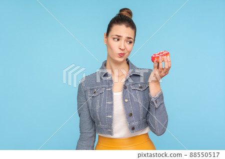 Unhealthy sweet food. Dissatisfied pensive woman with hair bun looking at donut with displeased expression and doubting to eat confectionary, junk food. indoor studio shot isolated on blue background 88550517