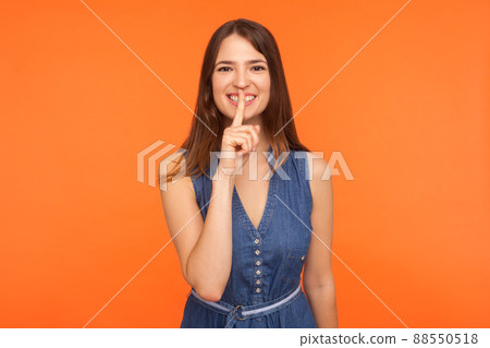 Please be quiet, don't speak. Positive brunette woman in denim dress making gesture to keep silence and smiling friendly, holding finger on lips, shushing hiding secret rumor. indoor, studio shot 88550518