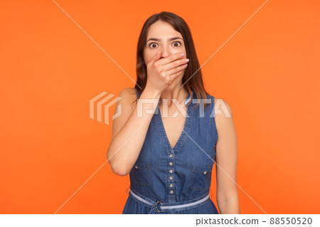 Scared brunette woman in denim dress covering mouth with hand and looking frightened at camera, afraid to talk, freedom of speech censorship concept. indoor studio shot isolated on orange background 88550520