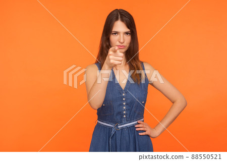 Hey you. Strict displeased brunette woman in denim dress pointing finger to camera and looking with suspicious angry expression, making accusing gesture. studio shot isolated on orange background Hey you. Strict displeased brunette woman in denim dress pointing finger to camera and looking with suspicious angry expression, making accusing gesture. studio shot isolated on orange background 88550521