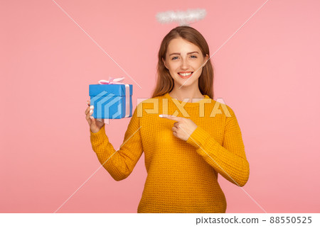 Look at my gift. Portrait of angelic beautiful ginger girl with halo over head pointing at present and smiling at camera, showing small wrapped box. indoor studio shot isolated on pink background 88550525
