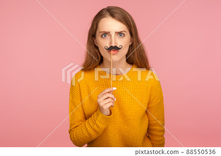 Portrait of nice looking charming ginger girl in sweater holding stick with curled mustache and sending kiss to camera, frowning looking bossy, masquerade. studio shot isolated on pink background 88550536