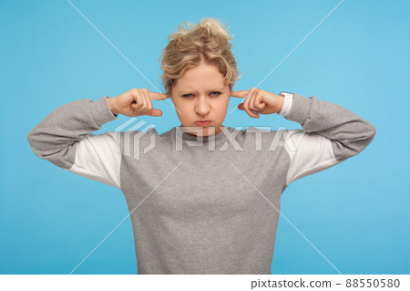 Don't want to hear. Irritated woman in grey sporty sweatshirt covering ears and puffing out cheeks, looking naughty and angry don't listen, ignoring annoying noise. indoor studio shot, blue background 88550580