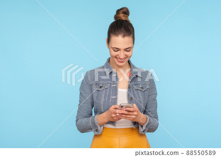 Portrait of pleased happy woman with hair bun in modern outfit typing on smartphone and smiling, reading funny message in social network, using mobile phone. studio shot isolated on blue background 88550589