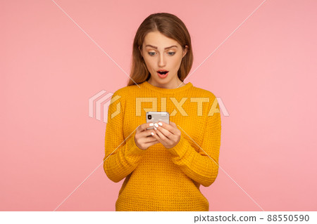 Portrait of surprised cute ginger girl texting on cellphone and expressing amazement, reading shocking message using smartphone, mobile application. indoor studio shot isolated on pink background 88550590