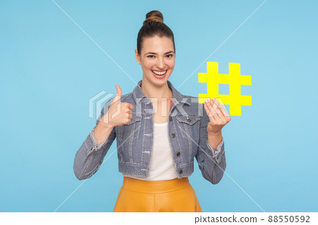 Like to popular blog and trends. Joyful attractive woman with hair bun in stylish outfit holding big yellow hashtag symbol and showing thumbs up gesture. indoor studio shot isolated on blue background Like to popular blog and trends. Joyful attractive woman with hair bun in stylish outfit holding big yellow hashtag symbol and showing thumbs up gesture. indoor studio shot isolated on blue background 88550592