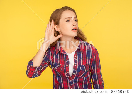 Can't hear you. Portrait of attentive ginger girl in checkered shirt holding hand near ear trying to listen quiet conversation, overhearing gossip. indoor studio shot isolated on yellow background 88550598