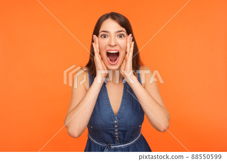 Oh my god, wow. Shocked impressed brunette woman in denim outfit standing with wide open mouth and clutching face in surprise, looking startled amazed at camera. indoor studio shot orange background 88550599