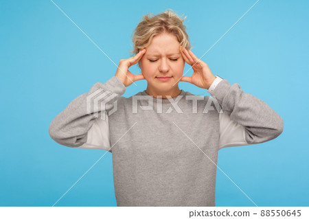 Portrait of depressed sick adult woman with short curly hair in sweatshirt holding head with hands, suffering headache, intense migraine of overwork. indoor studio shot isolated on blue background Portrait of depressed sick adult woman with short curly hair in sweatshirt holding head with hands, suffering headache, intense migraine of overwork. indoor studio shot isolated on blue background 88550645