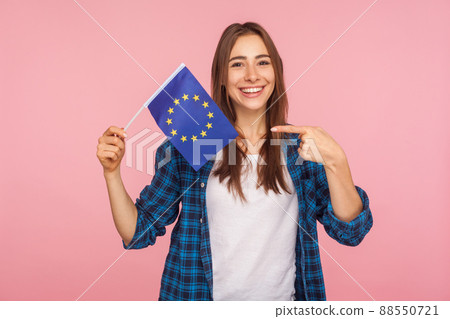 Portrait of positive friendly woman in checkered shirt smiling broadly and pointing at European Union flag, symbol of Europe, EU association, community. indoor studio shot isolated on pink background 88550721