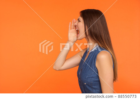 Side view of brunette woman in denim dress keeping arm near wide open mouth, screaming loudly, calling attention to important news, announcing event. indoor studio shot isolated on orange background 88550773