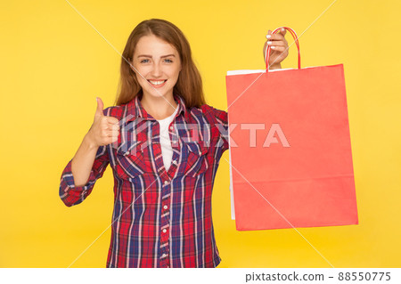 Pleased happy shopaholic, cheerful girl in shirt holding shopping bags and smiling at camera, showing thumbs up gesture, satisfied with purchase. indoor studio shot isolated on yellow background 88550775
