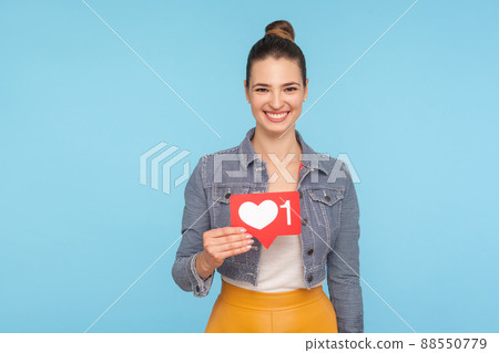 Portrait of joyful beautiful fashionably dressed woman with hair bun holding social media heart icon, network one Like button, follower notification symbol. studio shot isolated on blue background 88550779