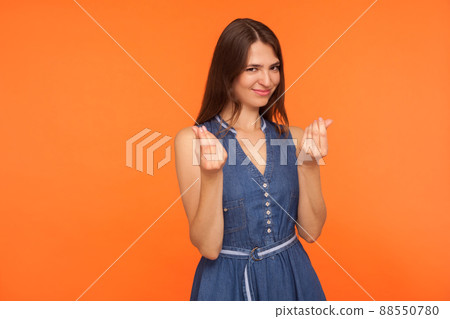 Attractive brunette woman in casual denim dress showing money gesture and looking slyly, having clever cunning plan how to make profitable business. indoor studio shot isolated on orange background 88550780