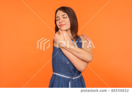 Self-love and egoism. Beautiful narcissistic brunette woman in denim dress embracing herself, feeling safe and comfortable, satisfied with appearance. indoor studio shot isolated on orange background Self-love and egoism. Beautiful narcissistic brunette woman in denim dress embracing herself, feeling safe and comfortable, satisfied with appearance. indoor studio shot isolated on orange background 88550832