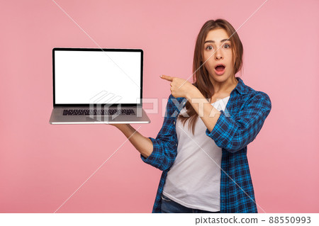 Portrait of amazed charming girl in checkered shirt showing blank laptop screen with shocked expression, pointing mock up, copy space for internet advertising. studio shot isolated on pink background 88550993