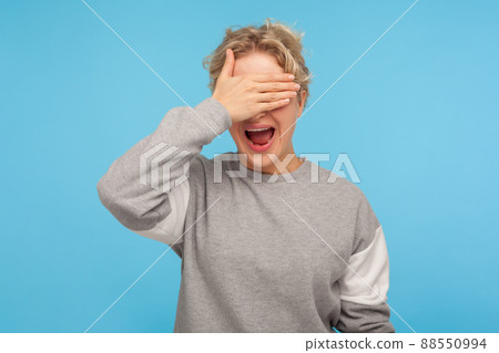 Don't want to look at this. Happy woman with curly hair in sweatshirt covering eyes and screaming with wide opened mouth, scared or ashamed to watch. indoor studio shot isolated on blue background 88550994