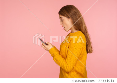 Side view of surprised beautiful ginger girl texting on cellphone and expressing amazement, reading shocking message using phone, mobile application. indoor studio shot isolated on pink background 88551013