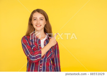 Attention to side. Portrait of cheerful ginger girl in casual shirt smiling and pointing to copy space, recommending to look at empty place, advertising. studio shot isolated on yellow background 88551029