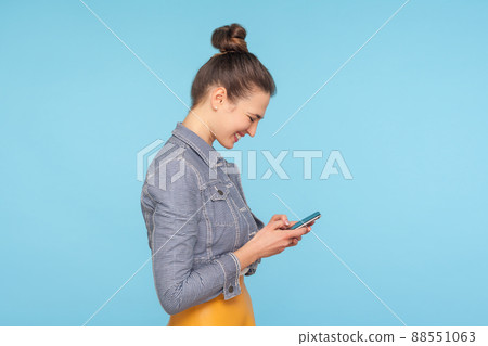 Side view of pleased cheerful woman with hair bun typing on smartphone and smiling, satisfied with mobile phone app, reading funny message in social network. studio shot isolated on blue background 88551063