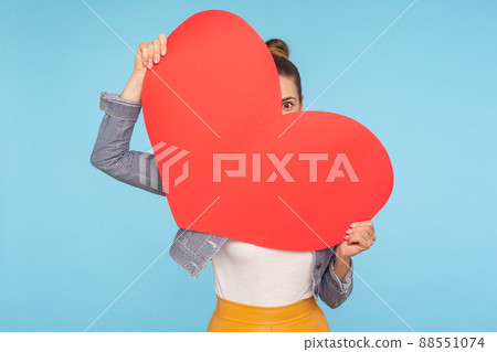 Portrait of funny optimistic woman peeking out of big red paper heart in her hands, looking with curious and amazed big eye, surprise on Valentines day. indoor studio shot isolated on blue background Portrait of funny optimistic woman peeking out of big red paper heart in her hands, looking with curious and amazed big eye, surprise on Valentines day. indoor studio shot isolated on blue background 88551074