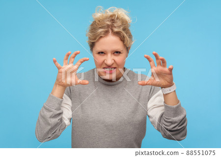Furious woman with curly hair in sweatshirt standing with raised claws and clenched teeth, experiencing strong anger, irritation, showing aggressive expression. indoor studio shot, blue background 88551075