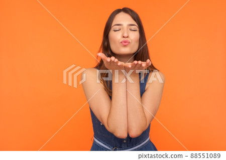 Cute romantic brunette woman standing with palms up and closed eyes in pleasure, sending amorous air kiss to camera, demonstrating affection, love. indoor studio shot isolated on orange background Cute romantic brunette woman standing with palms up and closed eyes in pleasure, sending amorous air kiss to camera, demonstrating affection, love. indoor studio shot isolated on orange background 88551089