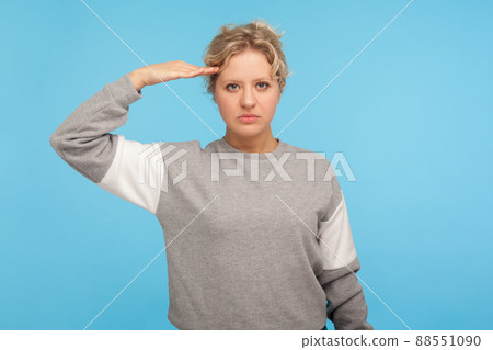 Yes sir. Serious confident adult woman with curly hair in sweatshirt looking at camera giving salute, listening to commander and ready to do order. indoor studio shot isolated on blue background 88551090