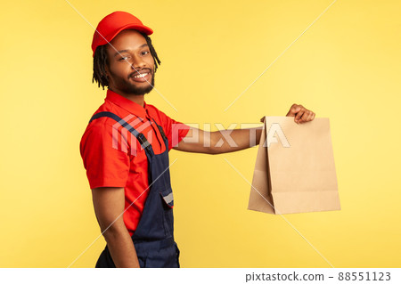 Side view portrait of happy professional courier wearing overalls giving groceries bag, delivering takeaway food, looking smiling to camera. Indoor studio shot isolated on yellow background. Side view portrait of happy professional courier wearing overalls giving groceries bag, delivering takeaway food, looking smiling to camera. Indoor studio shot isolated on yellow background. 88551123