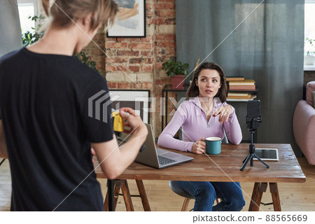 Attractive young woman with mug sitting at table with devices and discussing shooting plan with operator 88565669
