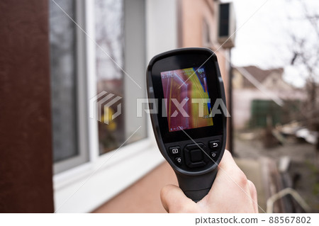 A male hand holds a thermal imager at the window of a house. Search for heat loss in private houses A male hand holds a thermal imager at the window of a house. Search for heat loss in private houses 88567802