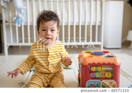 Baby boy playing with activity box in the bedroom while sitting on the floor wearing yellow pajama 88571320