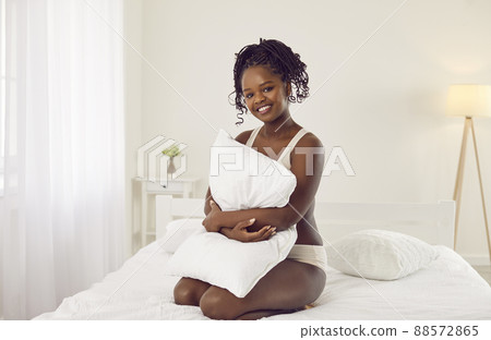 Happy beautiful young black woman sitting on bed at home and hugging soft white pillow Happy beautiful young black woman sitting on bed at home and hugging soft white pillow 88572865