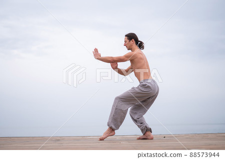 Caucasian man with naked torso practicing wushu on the seashore. Caucasian man with naked torso practicing wushu on the seashore. 88573944