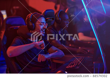 Happy young middle eastern professional gamer in headphones and eyeglasses dancing in front of computer while completing video game misson 88574302