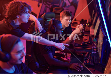 Young man pointing at computer screen while giving advice to African-American gamer focused on game in esports club Young man pointing at computer screen while giving advice to African-American gamer focused on game in esports club 88574340
