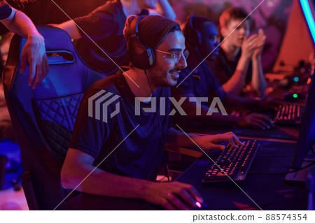 Smiling young professional gamer in hat and eyeglasses using computer while participating in esports tournament Smiling young professional gamer in hat and eyeglasses using computer while participating in esports tournament 88574354