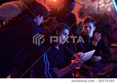 Young curly-haired man in headphones using tablet while explaining strategy of online game to friends in computer club Young curly-haired man in headphones using tablet while explaining strategy of online game to friends in computer club 88574369