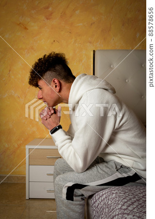 Caucasian young man with curly hair sitting on the bed with his hands on his chin thinking about the future that life holds for him, he is sad and melancholic due to the problems of acceptance. 88578656