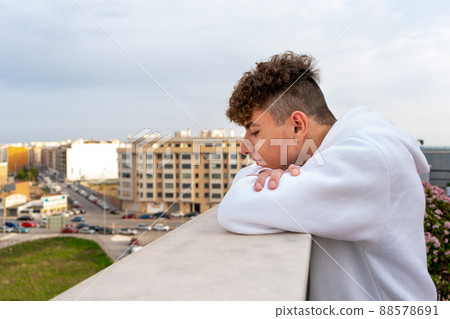 Young Caucasian man with curly hair dressed in a white sweatshirt looking at the city skyline serious and melancholic, I think about his future and what life has prepared for him 88578691