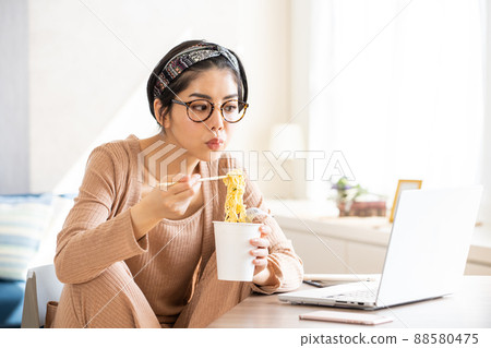 A young woman eating cup ramen while looking at her laptop 88580475