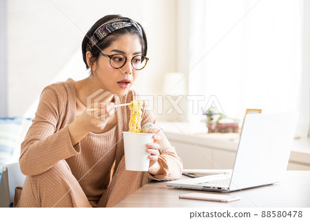 A young woman eating cup ramen while looking at her laptop A young woman eating cup ramen while looking at her laptop 88580478