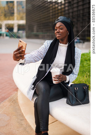 Smiling African American female wearing smart casual clothes and Muslim headscarf sitting with takeaway coffee and taking self portrait on cellphone in city Smiling African American female wearing smart casual clothes and Muslim headscarf sitting with takeaway coffee and taking self portrait on cellphone in city 88586675