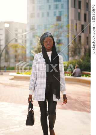 Gorgeous young African American female in classy outfit and Muslim headscarf with beret carrying handbag in city park and looking at camera Gorgeous young African American female in classy outfit and Muslim headscarf with beret carrying handbag in city park and looking at camera 88586676